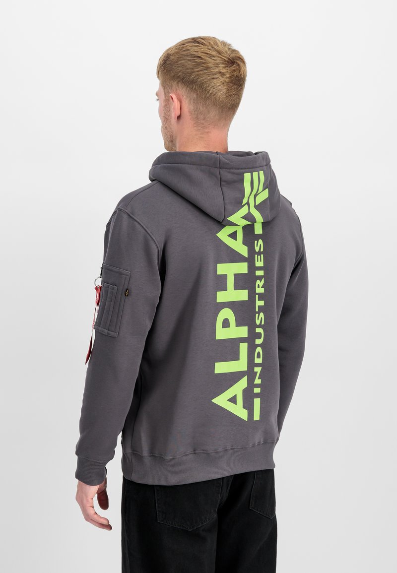 Mand med kort blondt hår iført en grå hættetrøje med lys grøn lodret "Alpha Industries" tekst på ryggen og sorte bukser.
