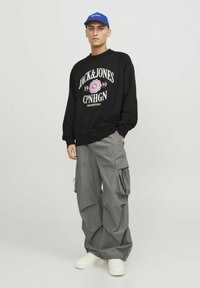 Zwarte sweatshirt met witte en roze grafische tekst; grijze cargobroek met grote zakken; witte sneakers; blauwe pet. Effen achtergrond.