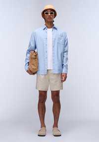 Camisa azul claro a rayas con botones, camiseta blanca, pantalones cortos beige, llevando un sombrero de cubo naranja y gafas de sol blancas, sosteniendo una bolsa beige.