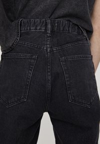 Jean noir taille haute, présentant une texture délavée, un design traditionnel à cinq poches, et des détails de couture visibles le long des coutures.