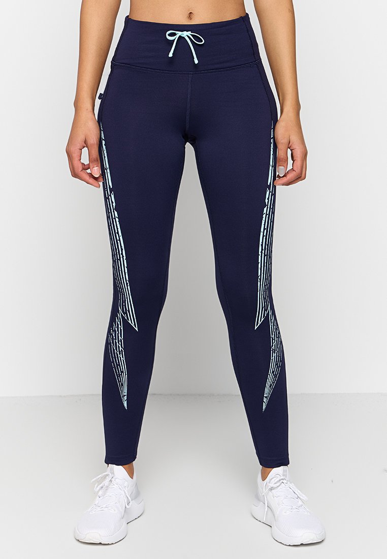 Mujer que lleva leggings azul marino con patrones geométricos en azul claro en los laterales y zapatillas deportivas blancas, de pie frente a un fondo liso.