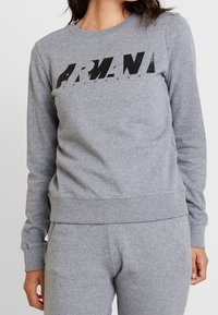 Sudadera gris de tejido suave con un gran logo negro "ARMANI EXCHANGE" en el pecho. Cuello redondo clásico y puños acanalados.