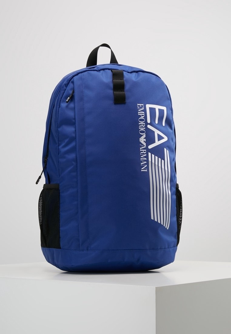 Sac à dos bleu Emporio Armani avec un grand logo blanc, des poches latérales en mesh noir et une poignée supérieure, présenté sur un socle blanc.