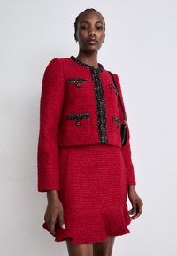 Rdeč tweed blazer z skrajšano silhueto, črnimi detajli in zlatimi gumbi. Usklajen z ujemajočo se teksturirano krilo z volani.
