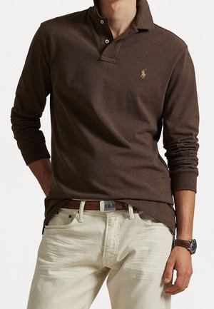 Man met een bruine poloshirt met lange mouwen, beige broek, een bruine riem en een polshorloge aan de linkerpols.