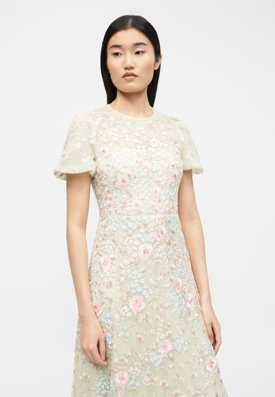 Γυναίκα με μακριά σκούρα μαλλιά που φοράει ανοιχτοπράσινο φόρεμα με διάφανα φουσκωτά μανίκια και ροζ και μπλε κεντήματα λουλουδιών.