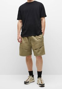 Zwarte t-shirt met korte mouwen gecombineerd met kaki cargoshorts met grote voorzakken. Model draagt zwarte sokken en beige sneakers.