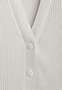 Gerippte, hellcremefarbene Strickjacke mit V-Ausschnitt und zwei weißen Knöpfen in der Mitte. Weiche Textur mit schlankem, figurbetontem Schnitt.