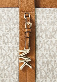 Tote en cuir marron avec un tissu blanc à motifs présentant le logo 'MK'. Accents en métal doré avec un emblème distinctif suspendu à la bandoulière.