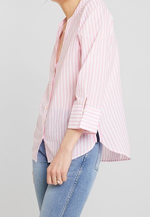 Kobieta ubrana w różowo-białą koszulę w pionowe paski, z podwiniętymi rękawami, oraz jasnoniebieskie dżinsy, stojąca na jednolitym tle.