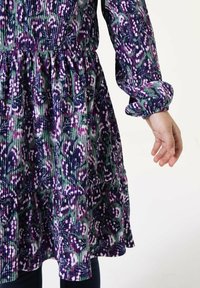 Lila und grün gemustertes Kleid aus gerippter Stoff mit langen Ärmeln, gerafftem Taillenbereich und Rüschensaum. Eine Hand ist von der Seite sichtbar.