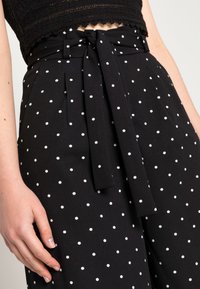 Svarta byxor med prickar i olika storlekar av vitt som är jämnt fördelade. Tillverkade av ett strukturerat tyg och har en knytning vid midjan.
