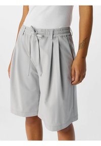 Shorts grigio chiaro con vita elastica e cordoncino regolabile. Presentano pieghe e una texture liscia, con una lunghezza che arriva appena sopra il ginocchio.
