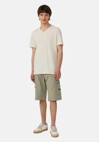 Een persoon draagt een crèmekleurig V-hals t-shirt, lichtgroene cargoshorts met zakken en beige sneakers met witte accenten. Eenvoudig ontwerp, casual pasvorm.