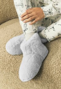 Harmaat tekoturkistossut, joissa pehmeä pinta, pyöreä varvas ja muhkea hihansuu, yhdistettynä kuvioituihin olohousuihin, joissa on rusetti- ja lasikuviot.