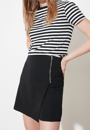 Femme portant une jupe noire avec une fermeture éclair sur le côté et une fente, associée à un haut à manches courtes rayé horizontalement noir et blanc.