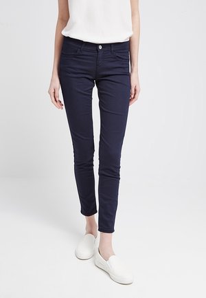 Femme portant un pantalon slim bleu foncé, un haut blanc sans manches et des chaussures blanches sans lacets, debout sur un fond clair.