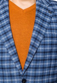 Niebieska marynarka w kratę o teksturowanym wykończeniu, z ciemnoniebieskimi i białymi wzorami w kratę, nałożona na pomarańczowy sweter z dekoltem w serek.