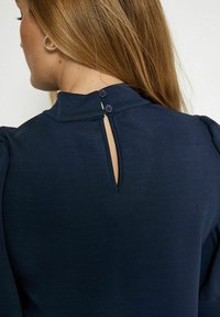 Dunkelblaues Oberteil mit hohem Kragen, Knopfdetails am Rücken und einer Schlüsselöffnung. Glatte Textur und weicher Stoff.