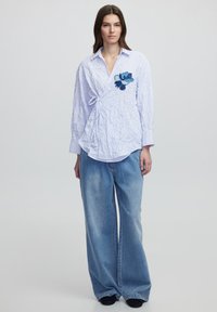 Camicia a righe bianca e blu con ricamo a fiore sulla sinistra, abbinata a jeans a palazzo in un lavaggio di denim chiaro.