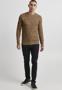 Hnědý pletený svetr s texturovaným povrchem, kulatým výstřihem a žebrovanými manžetami. Nosený přes černé slim-fit kalhoty a černé tenisky.
