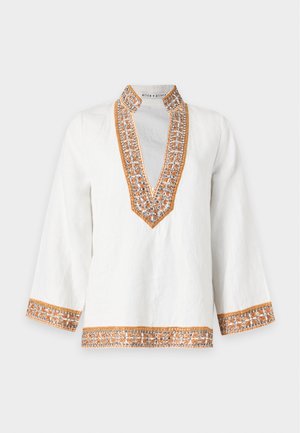 Femme portant une tunique blanche avec une bordure brodée orange, associée à un pantalon large foncé et des sandales à bout ouvert, debout devant un fond uni.