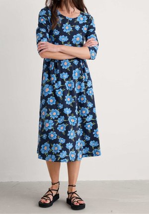 Kobieta ubrana w granatową sukienkę midi w niebieskie kwiatowe wzory, z rękawami do połowy przedramienia, w czarnych sandałach na platformie z paskami wokół kostek, stoi z założonymi ramionami.