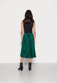 Vihreä hame, jossa on laskokset ja joka ulottuu polveen, yhdistetty mustaan ilmanhihaisella paidalla ja mustilla nilkkasaappailla. Sujuva pinta, korostetut laskokset, istuva vyötärö.