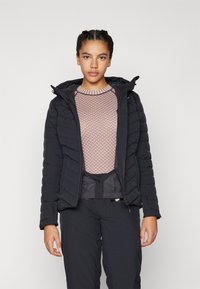Black puffer jacket with a textured surface, hood, and zip closure, worn over a light pink patterned sweater and black pants.