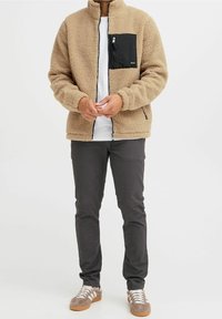 Giacca in pile beige con tasca sul petto nera con zip; presenta un colletto alto, due tasche laterali e una vestibilità rilassata.