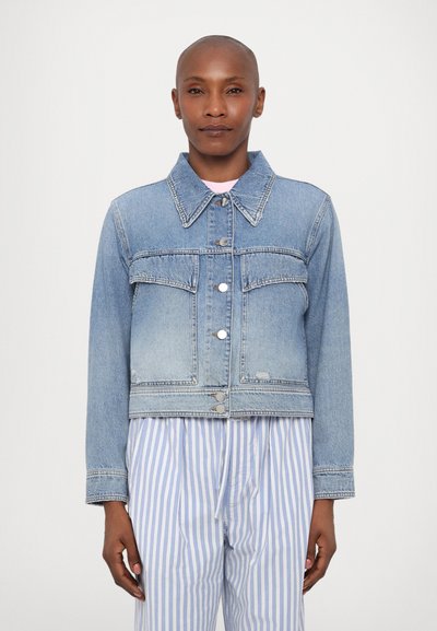 Chaqueta de mezclilla cortada en azul claro con cuello clásico, cierre de botones y dos bolsillos frontales. Lleva puesta sobre pantalones rayados en azul y blanco.