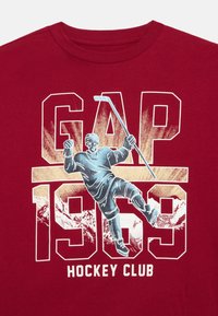 Piros póló, amelyen egy hoki játékos grafikus ábrázolása látható felszerelésben, ütővel a kezében, a "GAP 1969" és a "Hockey Club" felirat felett.