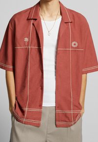 Camicetta a maniche corte color ruggine con ricami bianchi, caratterizzata da un motivo con palma e sole, con chiusura a bottoni e vestibilità rilassata.