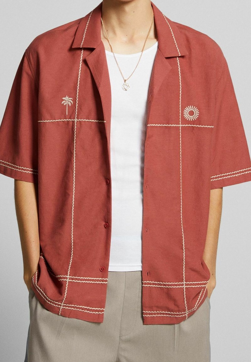 Camicetta a maniche corte color ruggine con ricami bianchi, caratterizzata da un motivo con palma e sole, con chiusura a bottoni e vestibilità rilassata.