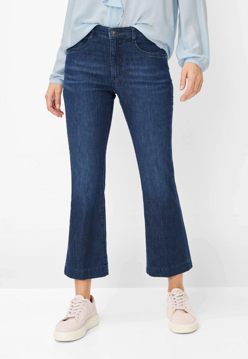 Kvinde iført korte mørkeblå flare-jeans, lyseblå bluse med lange ærmer og lyserøde snøresneakers, stående på hvidt gulv.