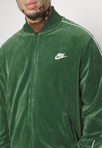 Veste Nike en velours vert avec fermeture éclair à l'avant, col côtelé et logo blanc. Comprend des poches latérales et une texture lisse.