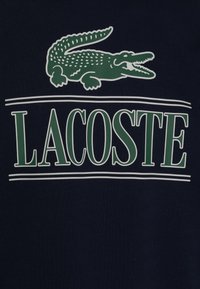 Temno modra tkanina z zelenim krokodilom nad besedilom "LACOSTE" v beli in zeleni barvi, vse v krepkem, preprostem tisku.