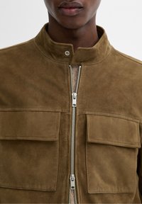 Giacca in suede marrone con colletto a cratere, zip argentata, due tasche sul petto e una finitura morbida e testurizzata.