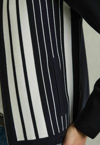Cardigan cu dungi bleumarin și albe, având dungi verticale, un buzunar lateral și un material texturat. Designul include un închidere cu fermoar.