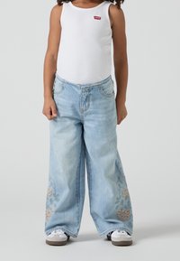 Světle modré kalhoty s širokými nohavicemi, na spodní části s květinovou výšivkou, v kombinaci s bílým ribbovaným tílkem. Obsahuje stříbrné kovové detaily.