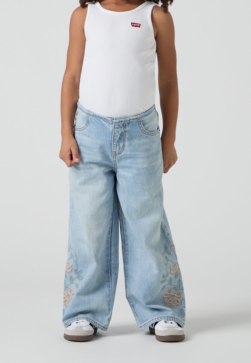 Světle modré kalhoty s širokými nohavicemi, na spodní části s květinovou výšivkou, v kombinaci s bílým ribbovaným tílkem. Obsahuje stříbrné kovové detaily.