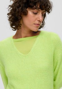 Limettengrüner, strukturiert gestrickter Pullover mit V-Ausschnitt, über einem glatten, hellgrünen Top getragen. Hat eine lockere Passform und weichen Stoff.