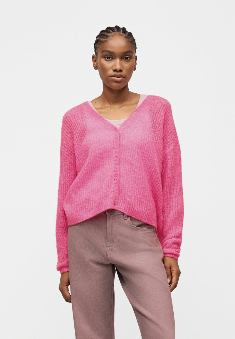 Γυναίκα με πλεξούδες, φορούν λαμπερή ροζ ζακέτα με κουμπιά και V-λαιμόκοψη πάνω από ένα ανοιχτό ροζ τοπ και μωβ παντελόνι, στέκεται μπροστά σε απλό φόντο.