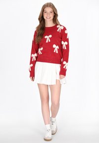 Maglione rosso lavorato a maglia con motivi di fiocchi bianchi e una leggera brillantezza, abbinato a una gonna bianca plissè e sneakers bianche.