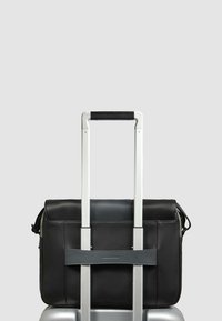 Schwarze Lederhandtasche, die an einem silbernen Koffergriff befestigt ist, mit glatter Textur, Klappenverschluss und einem grauen Riemen zur Befestigung.