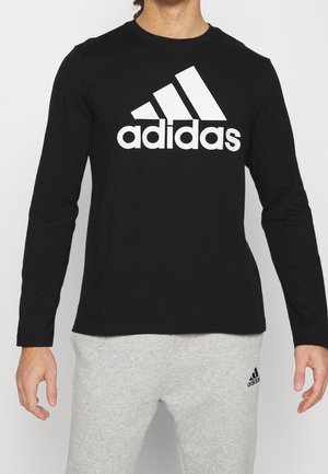 Osoba ubrana w czarną bluzkę z długim rękawem z dużym białym logo Adidas oraz jasnoszare spodnie dresowe z małym logo Adidas na lewej uda.