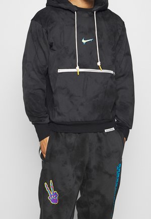 Man draagt een zwarte hoodie met witte trekkoorden en een blauw druipend logo, gecombineerd met een zwarte broek met kleurrijk skelet-hand vredesteken.