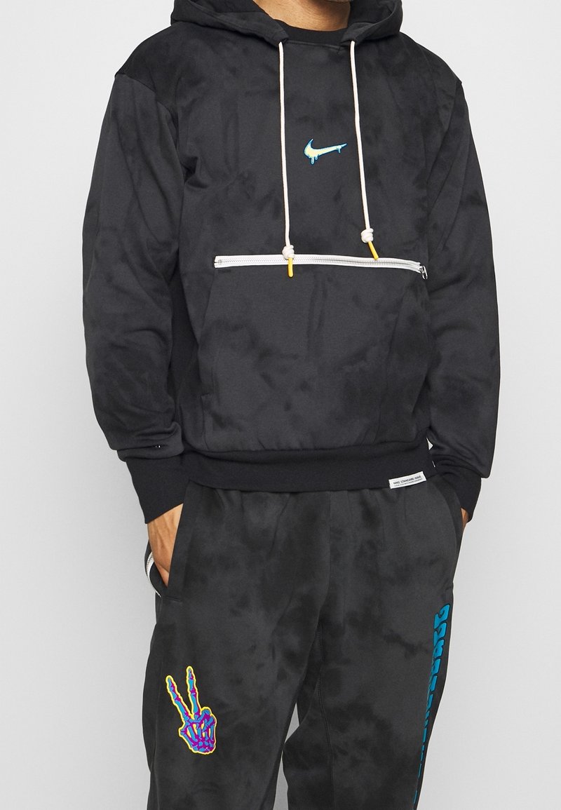 Homme portant un sweat à capuche noir avec des cordons blancs et un logo bleu dégoulinant, associé à un pantalon noir avec un signe de paix coloré en forme de main de squelette.