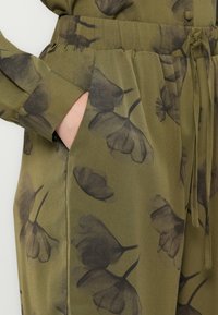 Primo piano di una mano nella tasca di pantaloni verde oliva con stampa floreale nera e una vita elastica con cordino.