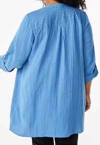 Chemise bleue légère avec un dos texturé et des détails œillets au niveau du bi-matière. Présente des manches longues retroussées et une coupe fluide et ample.